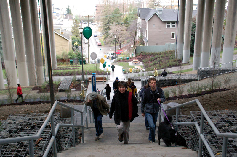People utilize stairs up a hillside under a freeway overpass.