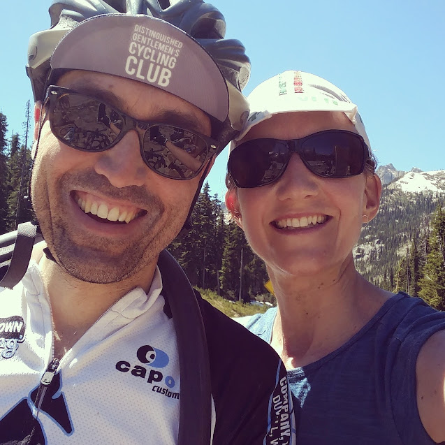 A person with a bike helmet and a person in a baseball hat smiling while in the mountains in the summertime.