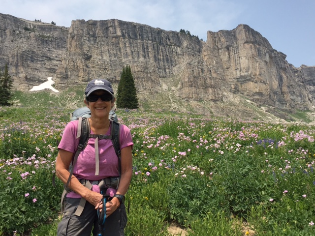 A hiker smiles at the camera while in a mountain meadow with sharp rock faces in the background.