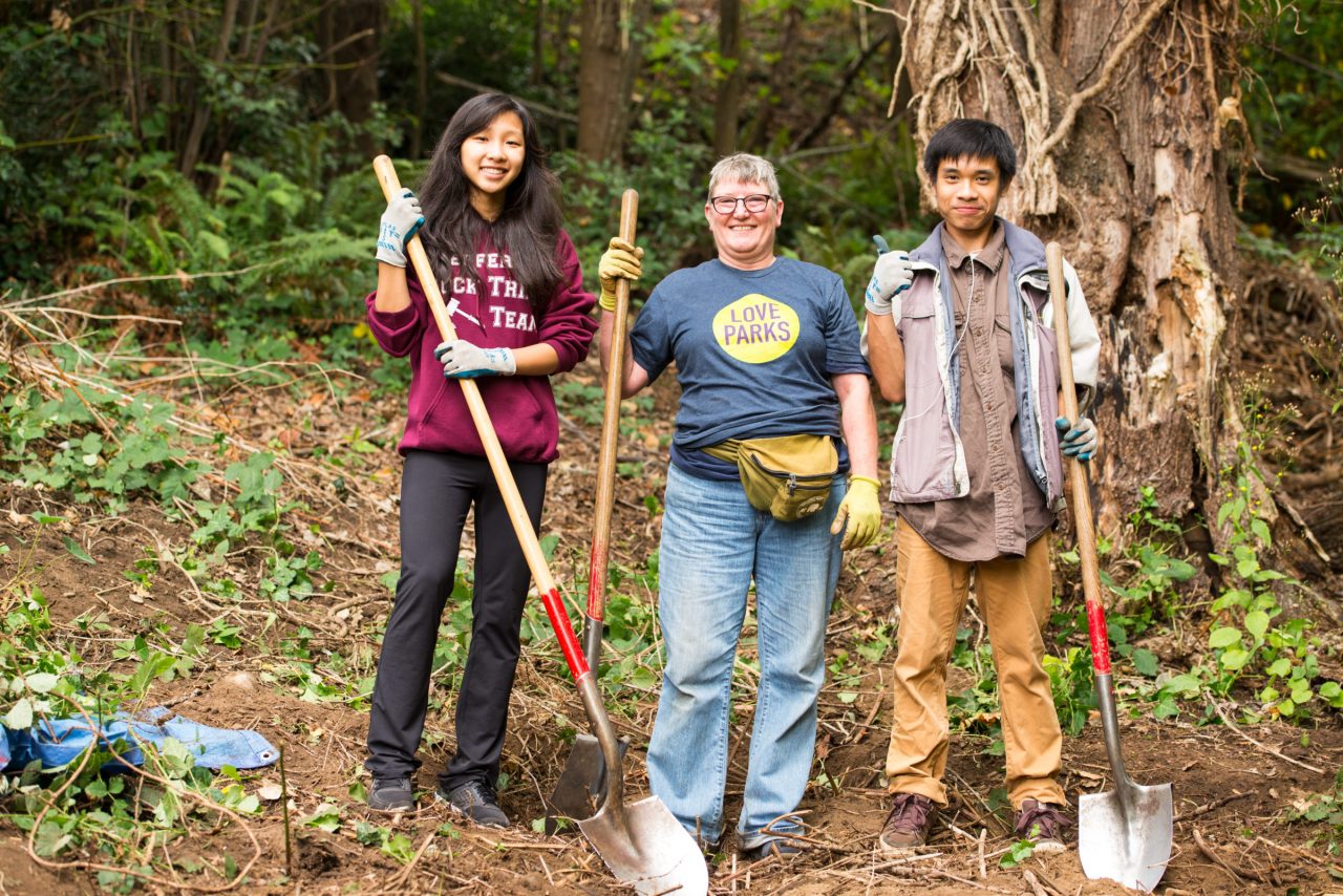 Two teens and an adult holding shovels and smiling towards the camera while working in the forest.